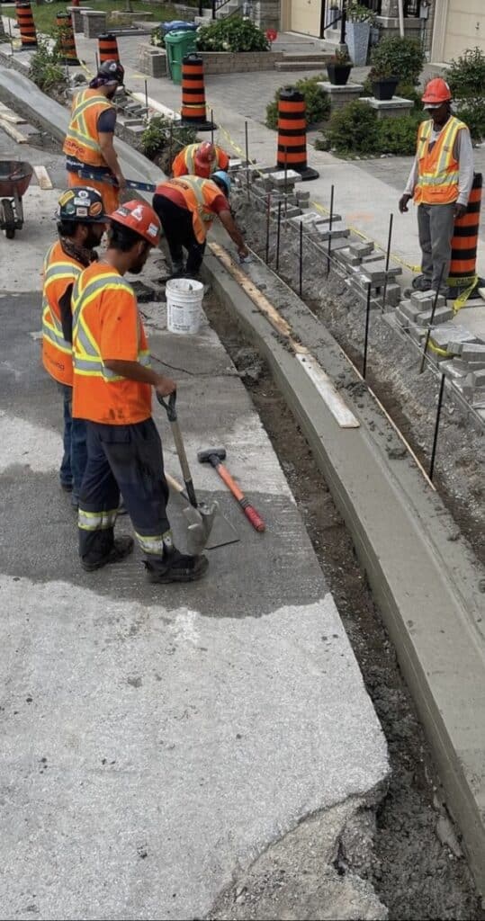 Construction workers in safety vests and helmets pouring and finishing asphalt on a road project, surrounded by construction materials and equipment, showcasing Strada Paving's expertise in asphalt paving and maintenance services.