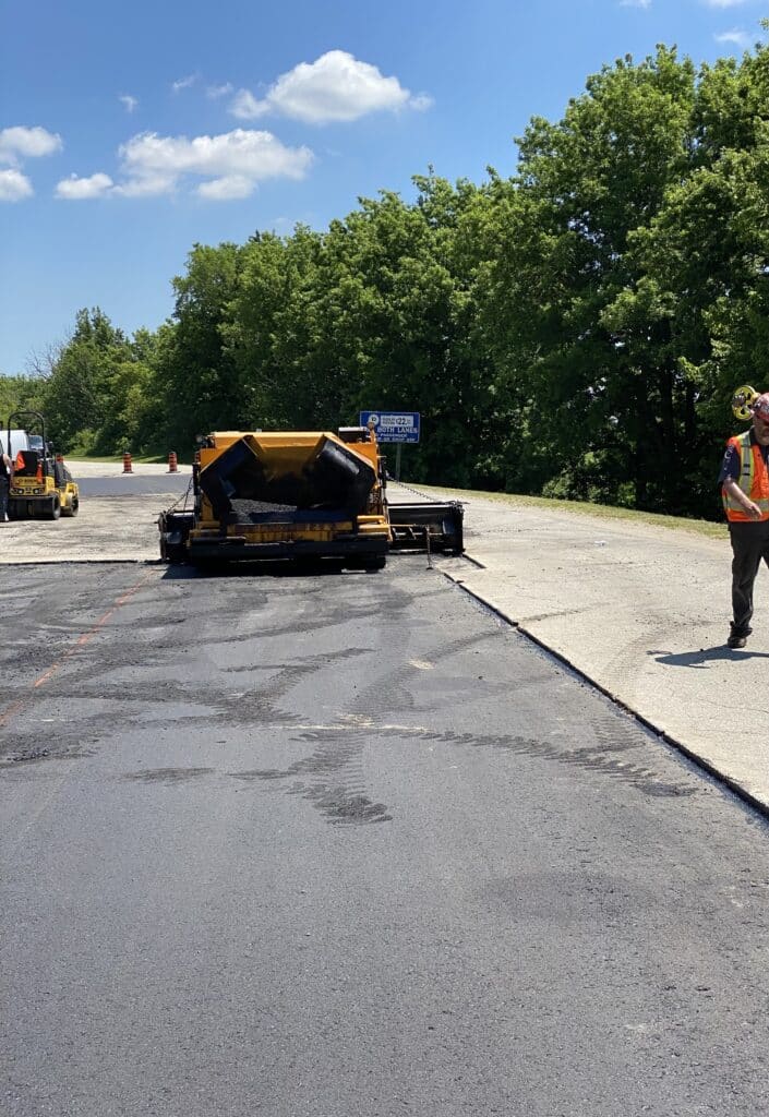 Asphalt paving machinery in operation on a newly paved surface, with a worker in safety gear overseeing the project, surrounded by trees and a clear blue sky, showcasing Strada Paving's expertise in asphalt paving services.