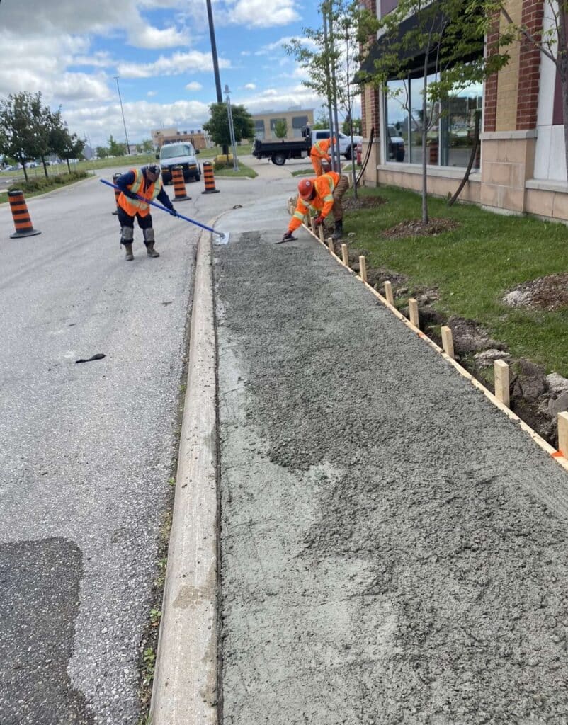 Workers in safety vests pouring and leveling fresh concrete for an asphalt paving project, with construction vehicles and traffic cones in the background.