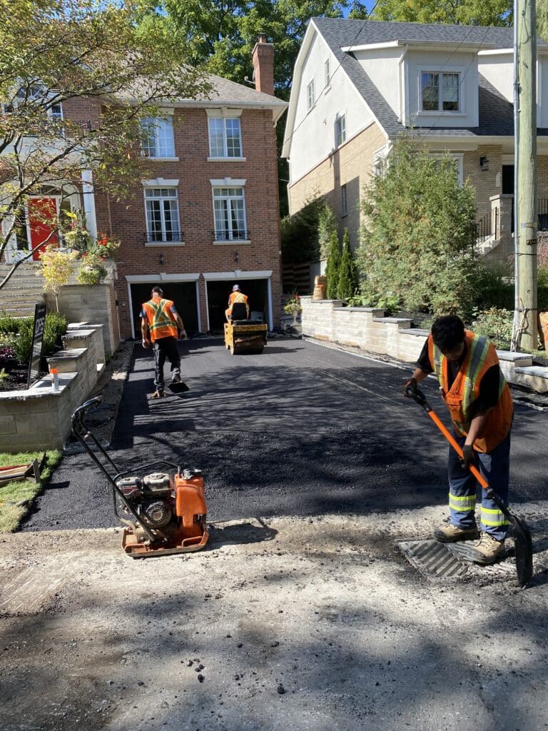 Workers in safety vests paving a residential driveway with asphalt, featuring a compactor and tools, showcasing Strada Paving's expertise in asphalt paving services in Toronto.