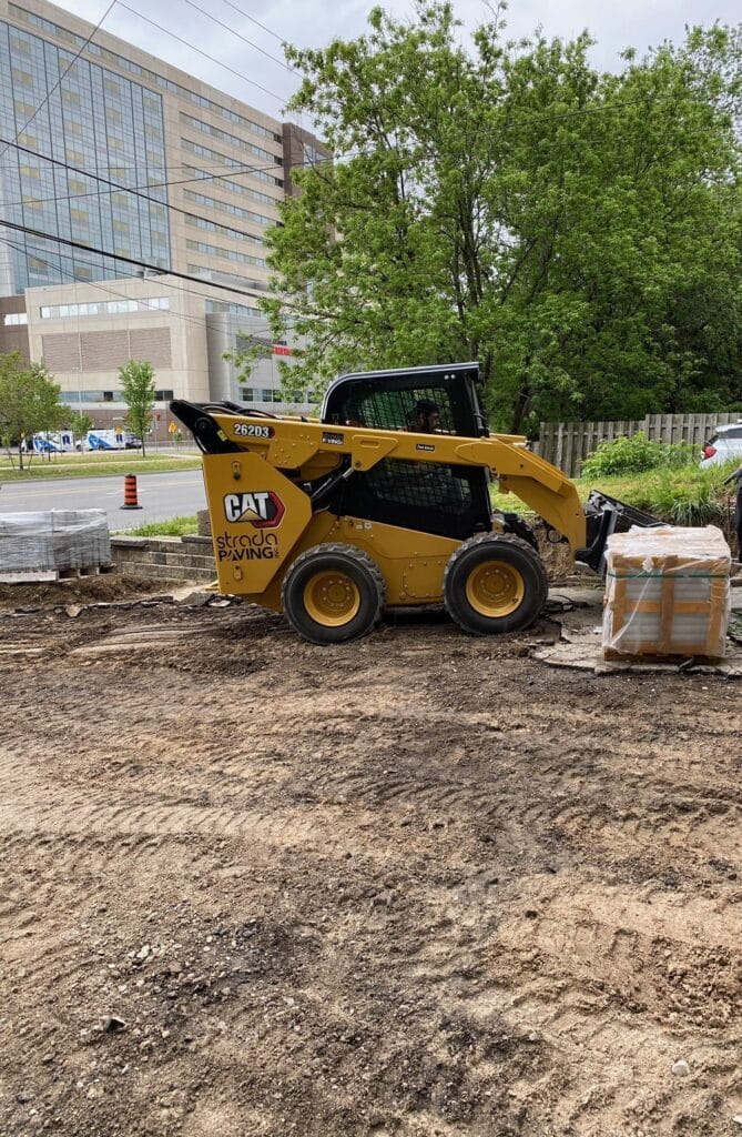 Yellow CAT skid steer loader with Strada Paving branding on construction site, surrounded by gravel and building in background, showcasing asphalt paving equipment.