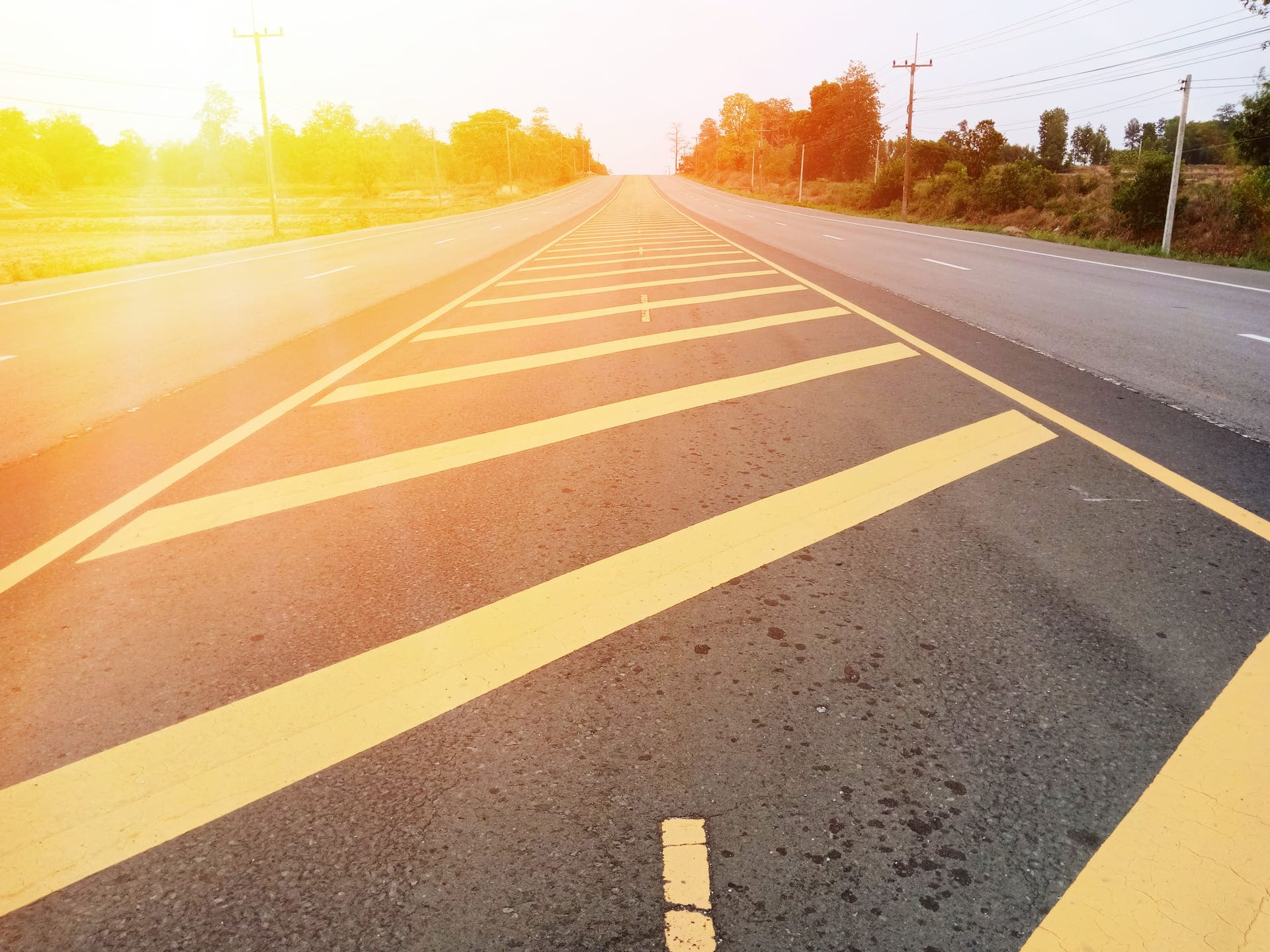 Freshly painted yellow diagonal pavement markings on an empty asphalt road during sunrise, indicating road lane guidance and safety regulations.