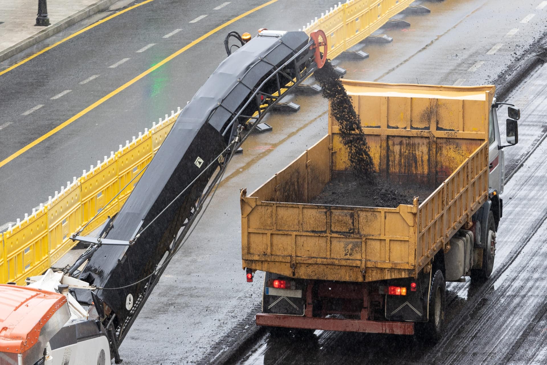 Asphalt milling machinery transferring milled asphalt into a yellow dump truck on a construction site, illustrating Strada Paving's asphalt milling services for road surface restoration.