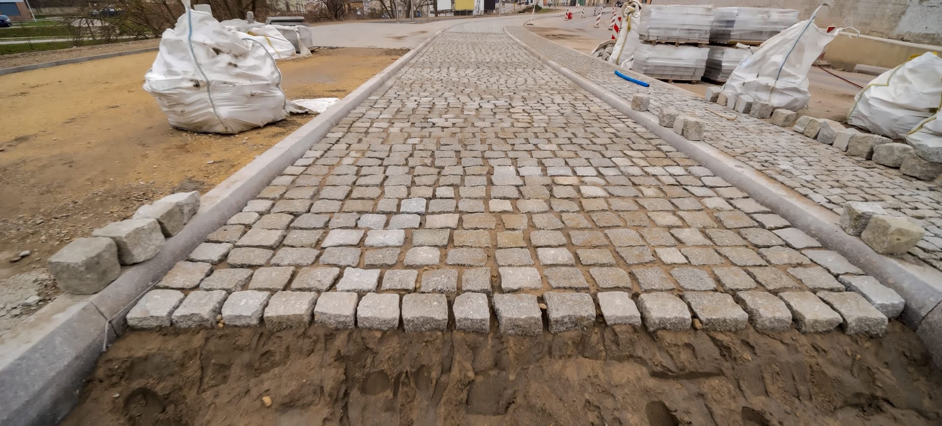 Paved concrete pathway under construction with cobblestones, surrounded by construction materials and bags, illustrating Strada Paving's durable concrete work for commercial and industrial surfaces.